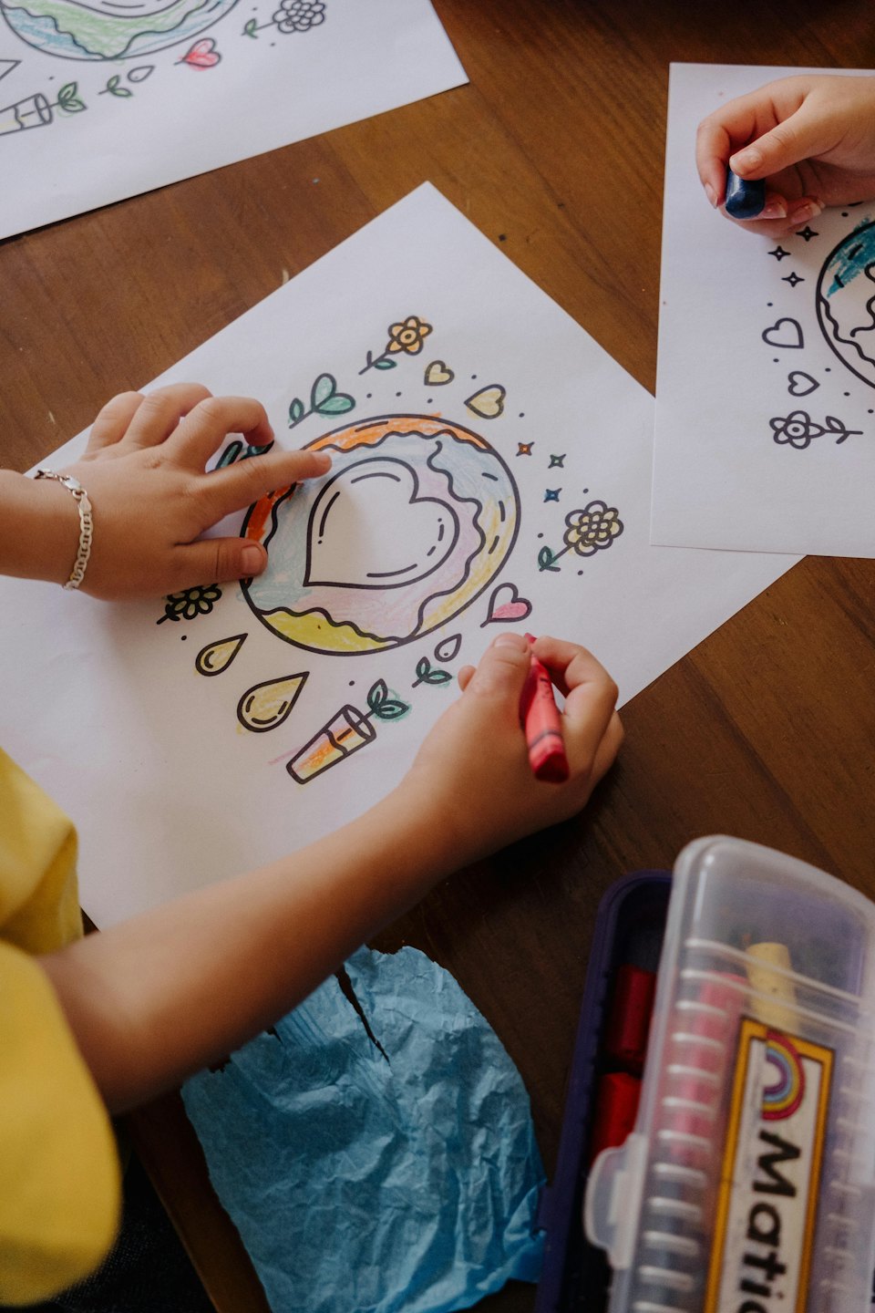 Children's hands coloring drawings with red and blue crayons.