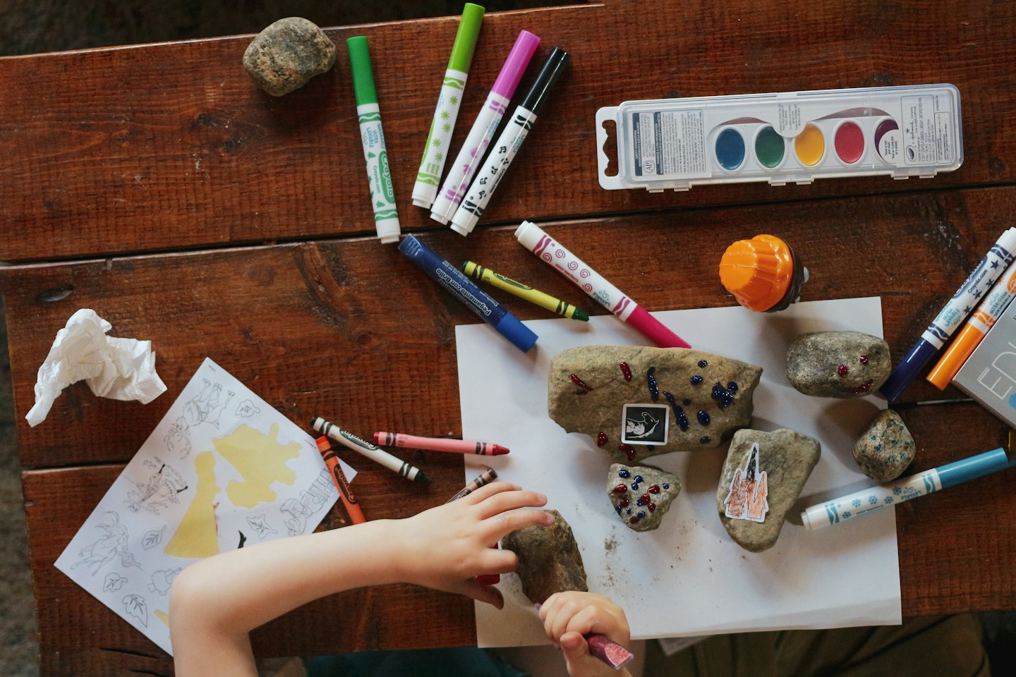 Child's hands decorating rocks with colorful paint on a wooden table.